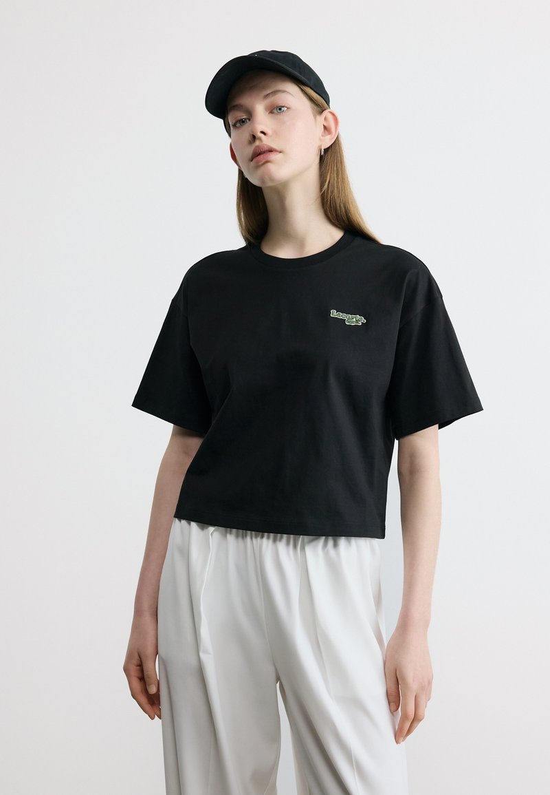 Νεαρή γυναίκα που φοράει μαύρο καπέλο, μαύρο κοντομάνικο μπλουζάκι με μικρό λογότυπο και φαρδιά λευκή παντελόνια μπροστά σε απλό φόντο.