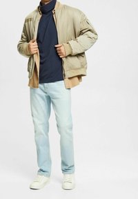 Beige bomberjacka med ljusbruna detaljer, marinblå polotröja, ljusblå slitna jeans och vita sneakers. Avslappnad passform, utan utsmyckningar.