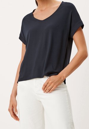 Femme portant un haut noir à manches courtes et décolleté en V, ample, avec un pantalon blanc, la main sur la hanche, sur un fond uni.