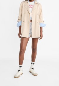 Manteau beige à double boutonnage avec manches retroussées, porté sur une chemise bleu clair et un short gris. Baskets blanches avec semelles beige clair, accents noirs, chaussettes rayées.