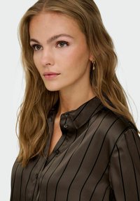 Blusa marrón a rayas hecha de tela suave, con cuello, botón en la parte delantera y mangas largas. Un sutil brillo acentúa la textura.