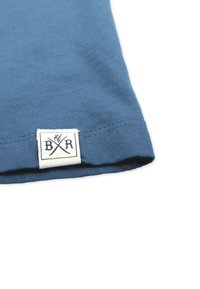 Blaues Baumwollgewebe mit einem umgeschlagenen Saum, das ein angenähtes Etikett mit einem gekreuzten Design und den Initialen "B/R" in Schwarz auf weißem Hintergrund präsentiert.