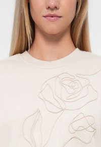 Beige sweatshirt met geborduurd rozenontwerp op de voorkant. Heeft een ronde hals en textuursteek voor extra detail.