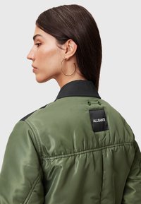 Grøn nylon bomberjakke med sort krave, har ribbet tekstur, elastiske manchetter og et ALLSAINTS mærke på bagsiden.