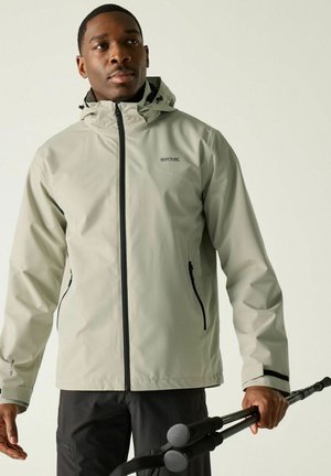 Hombre con chaqueta con capucha gris claro y pantalones negros, sosteniendo bastones de trekking negros plegados en la mano derecha.