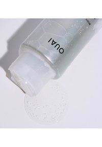 Transparent flaska med duschgel som har ett runt, vitt lock. Vätskan är ljusgrå, bubblig och har samlats på en vit yta i närheten. Varumärkeslogotypen är synlig.