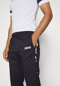 Zwarte sportbroeken met witte "JAKO" logo, elastische tailleband, zijzakken en contrasterende witte streepdetails. Textuur materiaal met een gladde afwerking.