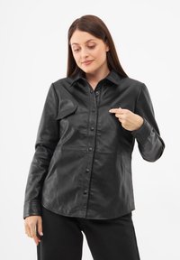 Giacca camicia in pelle nera caratterizzata da una chiusura frontale con bottoni, colletto e due tasche sul petto, con una silhouette aderente e una texture liscia.