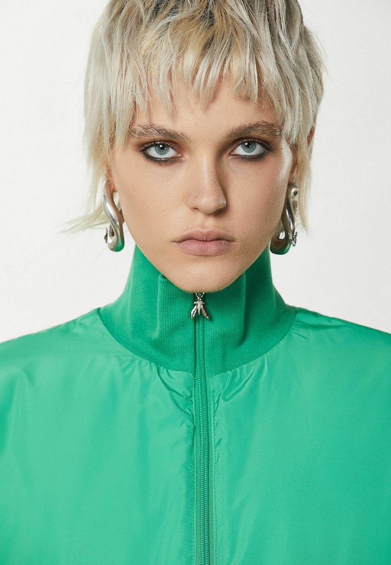 Zielona kurtka zapinana na zamek, z wysokim kołnierzem, wykonana z gładkiego, błyszczącego materiału. Posiada mały metalowy detal przy szyi oraz ogólny, elegancki wygląd.