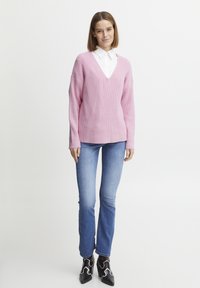 Roza pletena majica z V-izrezom in rebrasto teksturo, nošena čez belo srajco s kragnico, v kombinaciji s svetlo modrimi kavbojkami in črnimi škornji na peto.