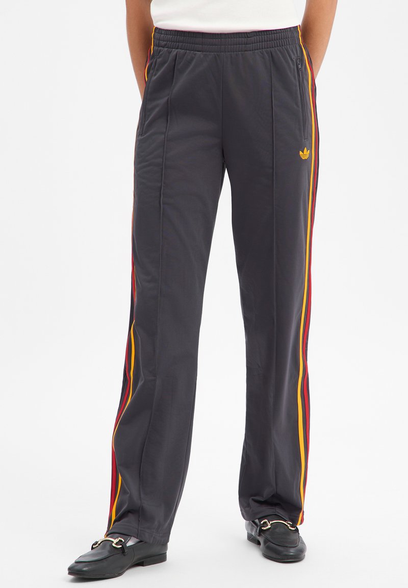 Personne portant un pantalon de survêtement Adidas noir avec des bandes rouges et jaunes sur les côtés et des mocassins noirs, debout les mains derrière le dos.