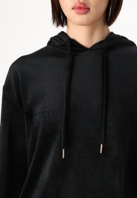 Sudadera negra hecha de un material suave y esponjoso, con un bolsillo tipo canguro en el frente, capucha con cordón y detalles metálicos en los extremos del cordón.