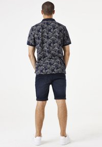 Marineblauwe polo-shirt met een bladerenpatroon in lichtgrijs, korte mouwen en een kraag; gecombineerd met opgerolde denim shorts en witte sneakers.