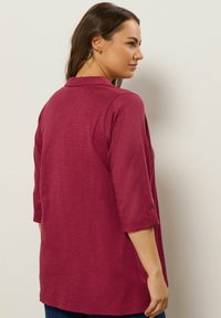 Blusa bordeaux con colletto, maniche a tre quarti e bottoni decorativi ai polsini. Tessuto liscio, vestibilità leggermente ampia.