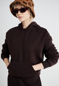 Sweatshirt capuz castanho feito de tecido suave, com um bolso frontal tipo canguru e punhos canelados. Usado com um chapéu de balde castanho estampado.