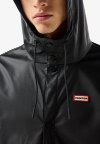 Közeli kép egy személyről, aki fekete kapucnis esőkabátot visel húzózsinórokkal és a mellkasán egy piros szegélyű Hunter logóval ellátott felvarróval.