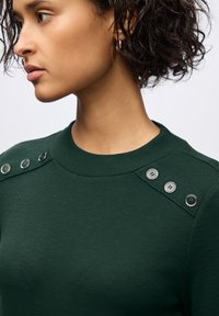Top a coste verde scuro con scollatura a crew e bottoni decorativi argentati sulla spalla. Texture liscia con una forma aderente.