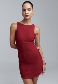 Punane vormi istuv kleit tekstuurse pinnaga, varrukateta disain, ümara kaelusega ja määratletud vööjoonega. Kulla toonis kõrvarõngad lisavad detaili.