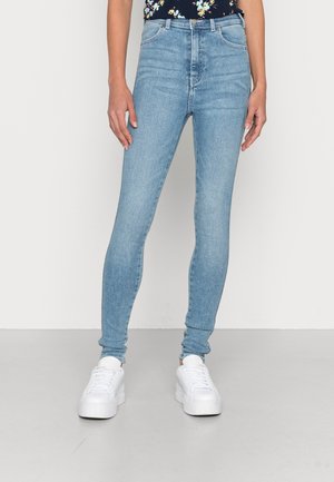 Femme portant un jean skinny bleu clair et des baskets à plateforme blanches, debout devant un fond clair uni.