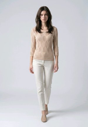 Mujer de pie frente a un fondo liso, vistiendo un suéter de punto beige con estampado, pantalones color crema y zapatos tipo slip-on en tono marrón claro.
