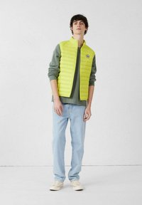 Limoengroene gewatteerde bodywarmer met hoge kraag, gedragen over een grijze trui, gecombineerd met lichtblauwe spijkerbroek en beige sneakers. Simpel ontwerp.