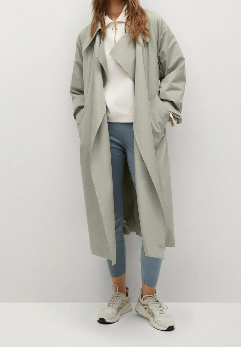 Femme portant un long trench-coat gris clair, un pull blanc zippé, un pantalon slim bleu et des baskets beige, debout devant un fond clair uni.