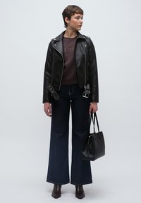 Fekete bőr motoros dzseki barna szöveges pulóverrel, sötétkék bővülő farmernadrággal és magassarkú bokacsizmával párosítva. Egy kis fekete táskát tart a kezében.