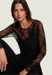 Top negro transparente con mangas largas de encaje, patrones florales y escote redondo, combinado con una prenda inferior negra. Modelo sentada, mostrando un anillo.