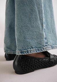 Zapatos planos tejidos negros con un respaldo de cuero suave, combinados con pantalones de mezclilla de pierna ancha en un tono azul claro con un dobladillo deshilachado, mostrando un tejido texturizado.