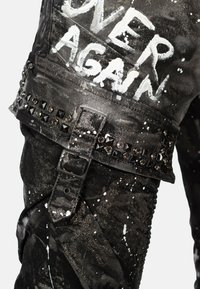 Pantalons en denim noir avec des éclaboussures de peinture blanche, des accents cloutés et une boucle décorative sur la taille avec "OVER AGAIN" imprimé en blanc.