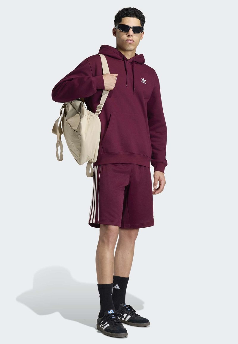 Giovane uomo che indossa felpa con cappuccio e pantaloncini Adidas color bordeaux, sneakers nere Adidas e calzini, occhiali da sole, con zaino beige sulla spalla.