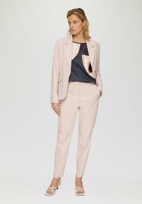 Lichtroest op maat gemaakte pak met een blazer en aansluitende broek, gecombineerd met een donkere blouse met abstracte patronen en accenten. Witte sandalen met bandjes.