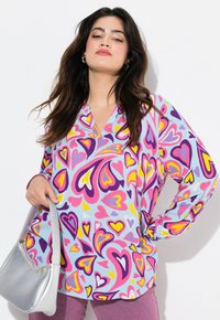 Blusa colorida de manga larga en tela azul claro con un patrón de corazones en rosa, púrpura y amarillo; presenta un escote en V y un corte holgado.