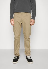 Beige katoenen broek met een recht model, voorzien van een knoopsluiting en voorzakken, gedragen met zwart-witte sneakers.