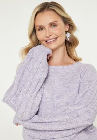Maglione lavorato a maglia di un leggero colore lavanda con maniche testurizzate a coste e scollatura a scallop. Il tessuto sembra morbido e peloso, creando un aspetto accogliente.