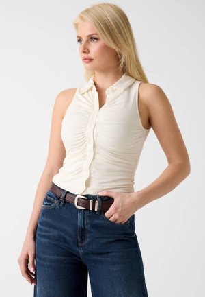 Blonde woman wearing sleeveless white ruched blouse with collar tucked into high-waisted dark blue jeans and a brown leather belt.