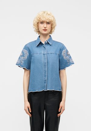 Person iført en kortærmet denimskjorte med hvid blomsterbroderi på ærmerne og bølgede kanter, kombineret med sorte bukser.