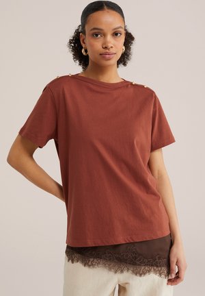 Jeune femme aux cheveux bouclés portant un haut manches courtes couleur rouille avec des boutons dorés sur les épaules et un pantalon beige avec un bord en dentelle.