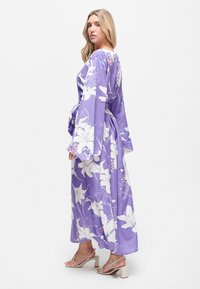Lila Blumen-Maxikleid aus leichtem Stoff mit langen Ärmeln und einem Taillenzug. Enthält weiße Lilien und pinke Akzente. Sichtbare Absatzsandalen.