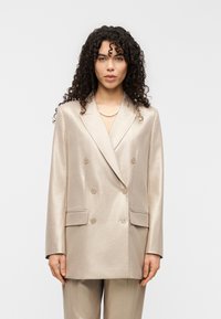 Διπλόμπουτο σακάκι σε μεταλλικό μπεζ ύφασμα με δύο εμπρόσθιες τσέπες, έξι κουμπιά και εφαρμοστή γραμμή, συμπληρωμένο με παντελόνι που ταιριάζει.