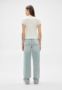 Sieviete ar gariem tumšiem matiem, valkā baltu īsrokas krekliņu un gaiši zilas platajām biksēm, stāvot ar muguru pret pilnīgi vienkāršu fonu.