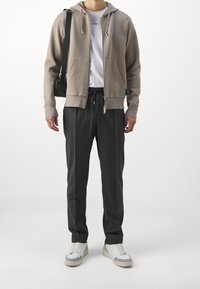 Beige Kapuzenpullover mit Fronttaschen, kombiniert mit dunkelgrauen Hosen. Weiße Sneakers mit grauen Akzenten runden das Outfit ab.
