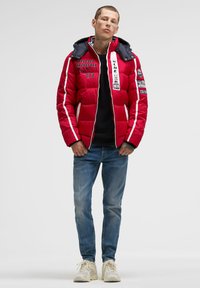 Red puffer jacket with black hood, white stripes, and multi-colored patches. Paired with blue jeans and beige sneakers.