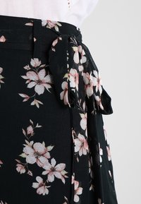 Černá květinová zavinovací sukně s růžovými a bílými květinovými vzory, texturovaná tkanina, s vázaným opaskem v pase.