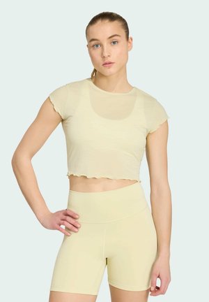 Vrouw met een lichte huid en blauwe ogen die een lichtgeel cropped T-shirt en hoge taille biker shorts draagt, staand met één hand op de heup.