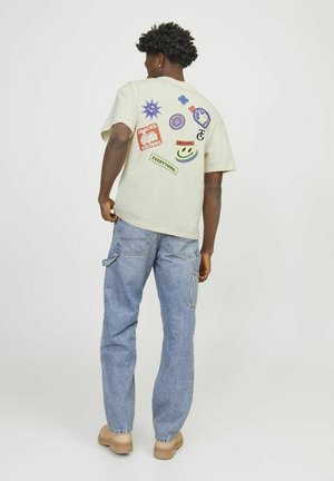 Camiseta de un color beige claro con pegatinas gráficas coloridas en la parte de atrás, combinada con unos jeans de mezclilla azules y zapatillas beige.