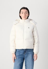 Hvit pufferjakke med høy krage, glidelås og trykknapper. Har et brodert logo og elastiske mansjetter. Brukt over blå jeans.