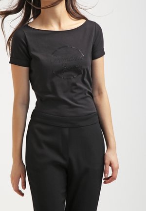 Femme portant un haut noir ajusté à manches courtes avec revers roulés et un pantalon noir assorti à taille haute, sur un fond clair.