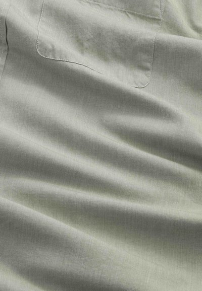 Hellgrauer Stoff mit einer aufgenähten quadratischen Tasche und sichtbarer Textur, die weiche Falten und Schatten zeigt.