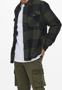 Chaqueta camisa de franela a cuadros verde y negro con dos bolsillos delanteros, cierre de botones y tela texturizada. Combinada con pantalones cargo oliva.